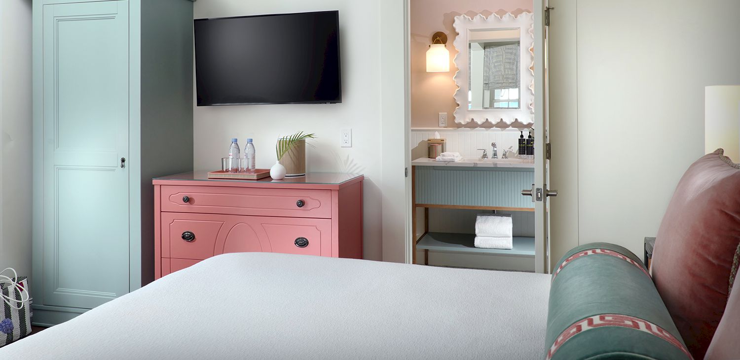 Modern hotel bedroom with a wall-mounted TV, a pink dresser, and a vanity mirror opening with towels and toiletries.