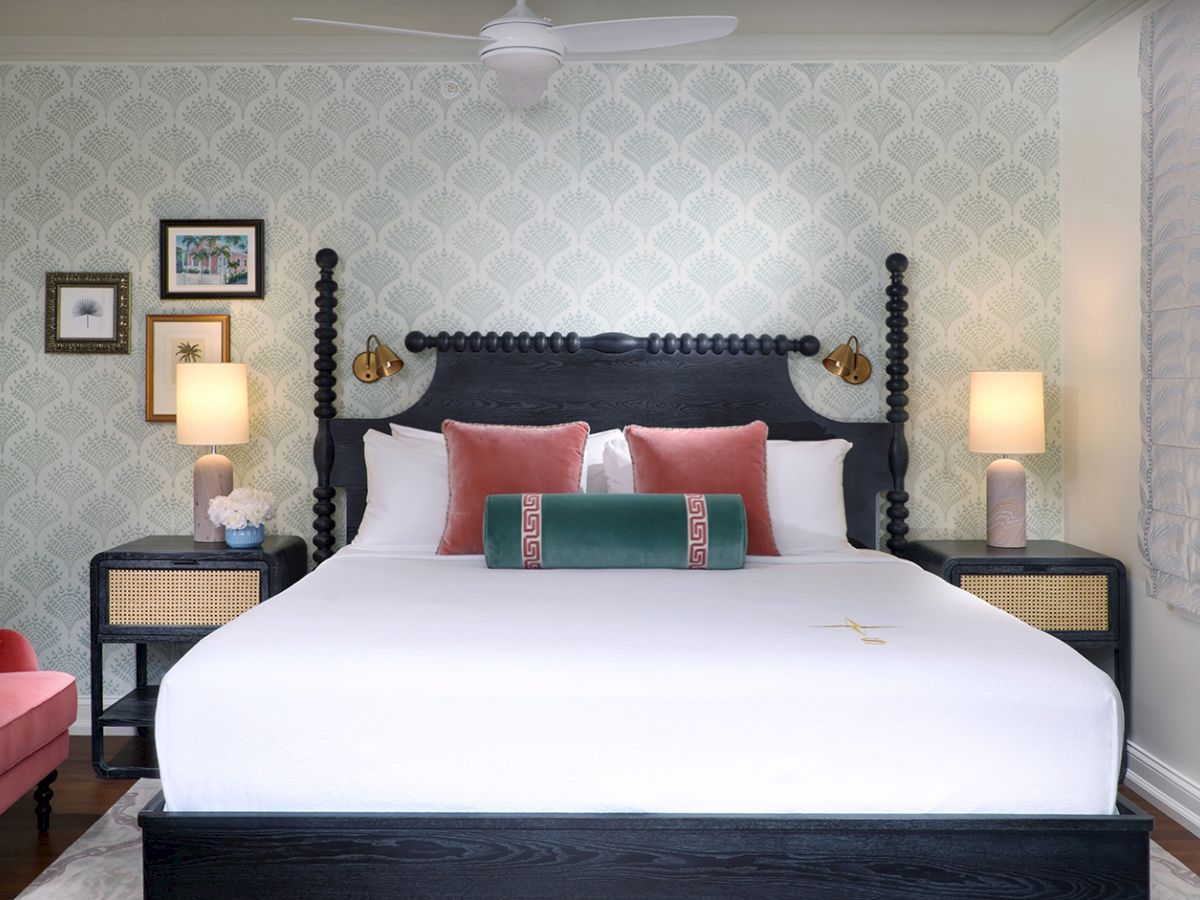 A bright, elegant bedroom with a dark four-poster bed, white linens, coral and teal pillows, lamps, framed art, and patterned walls.