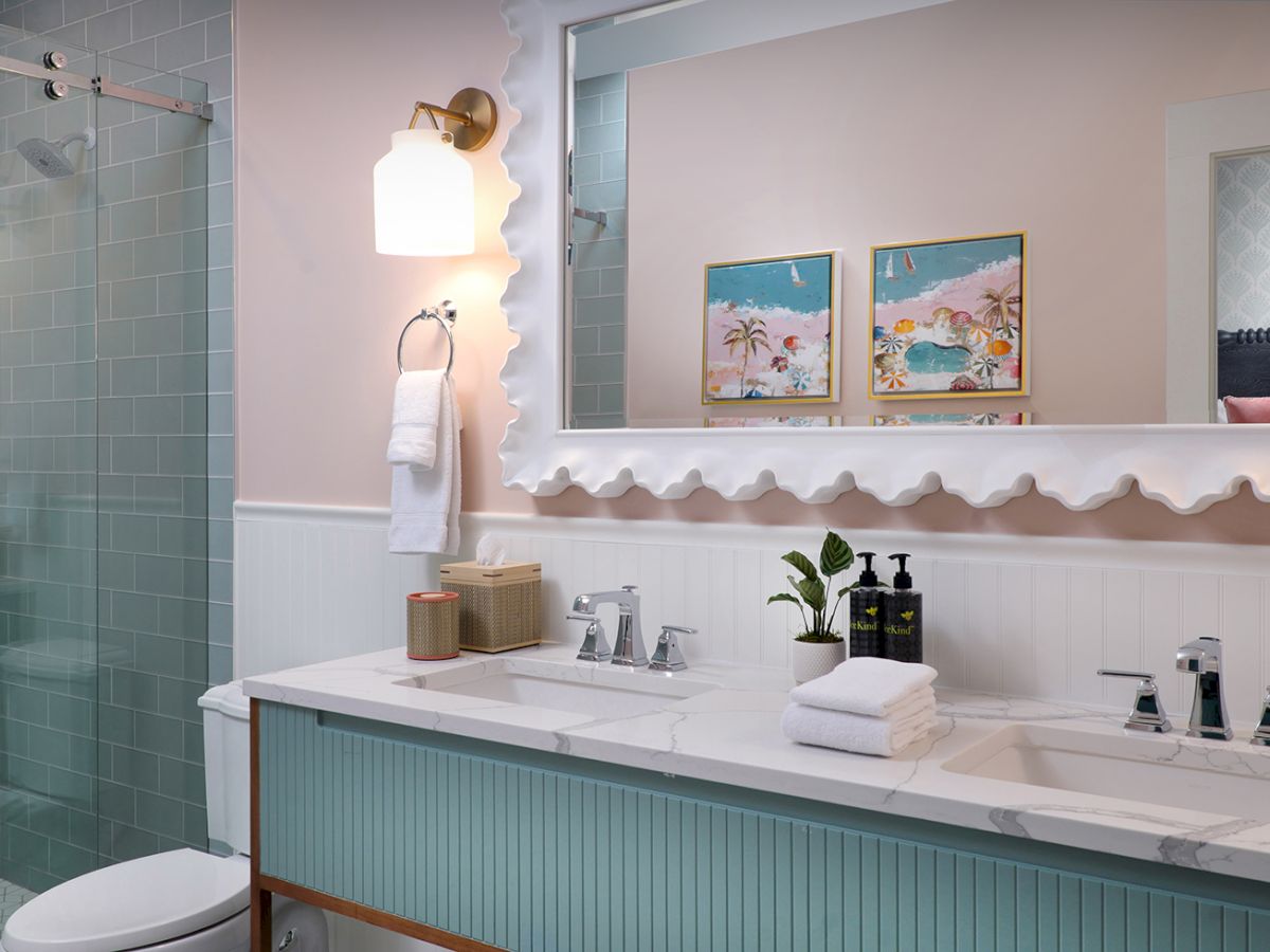 A bright modern bathroom with a marble vanity, chrome faucets, framed beach art, a towel, and a glass shower stall.