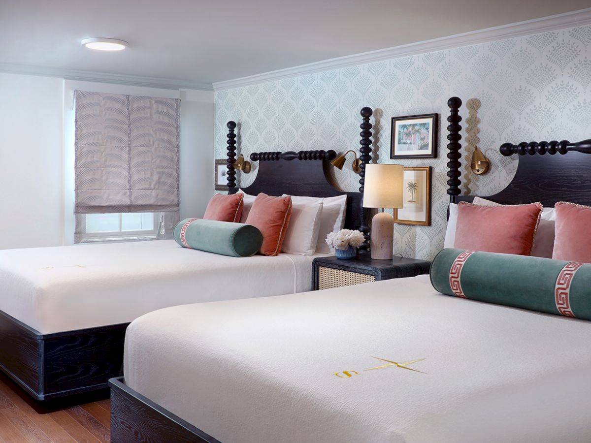 A bright modern bedroom with two large beds, patterned wallpaper, decorative pillows, and bedside lamps.