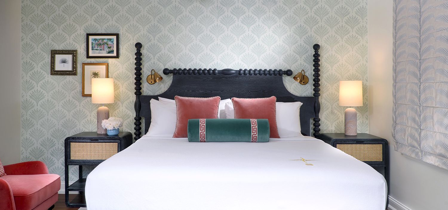 A stylish hotel bedroom with a dark four-poster bed, white linens, red pillows, bedside lamps, and patterned wallpaper.