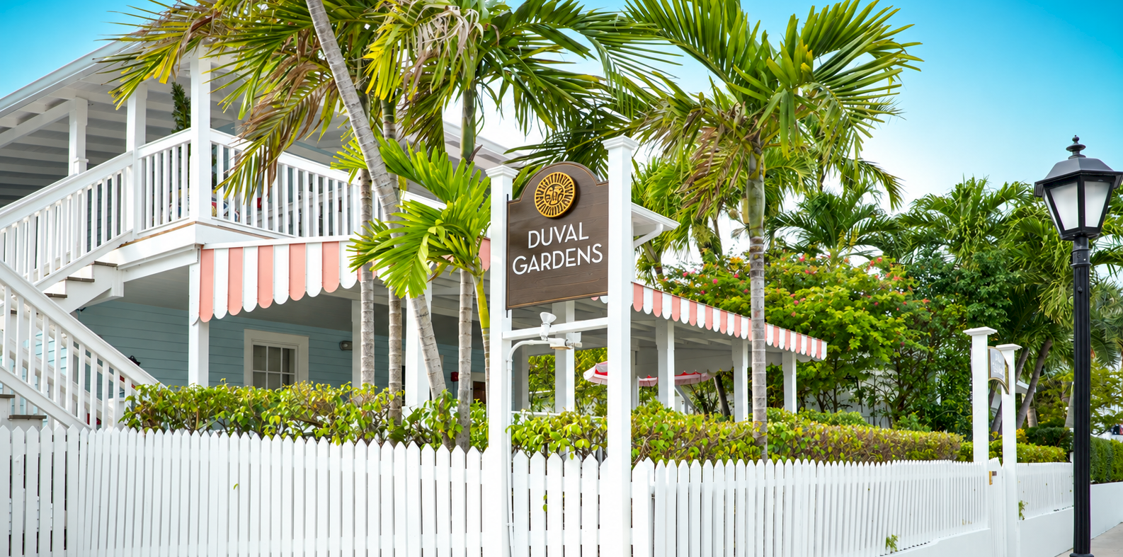 A sunny garden walkway outside Duval Gardens, with white railings, pink awning, and tall palm trees.