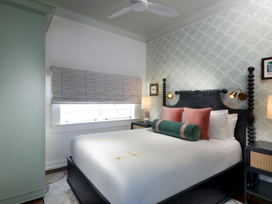 A cozy bedroom with patterned wallpaper, a dark wooden bed, layered pillows, bedside lamps, and a window with blinds.