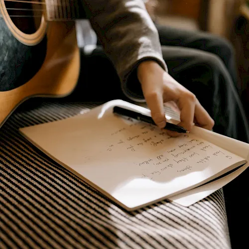 A person with an acoustic guitar scribbles notes on sheet music while holding a pen.