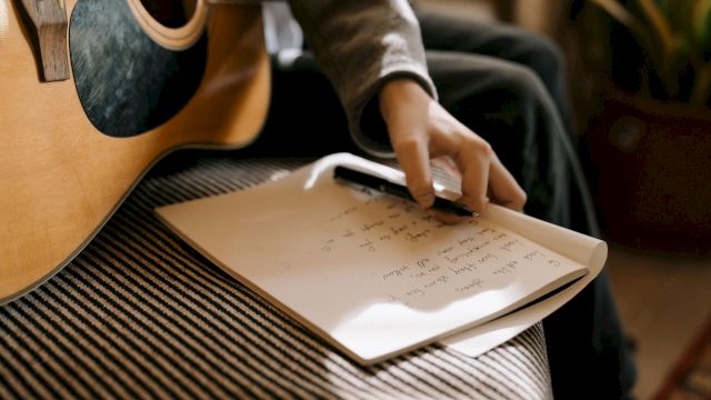A person in a cozy room writes notes on paper with a pen while a guitar sits nearby.