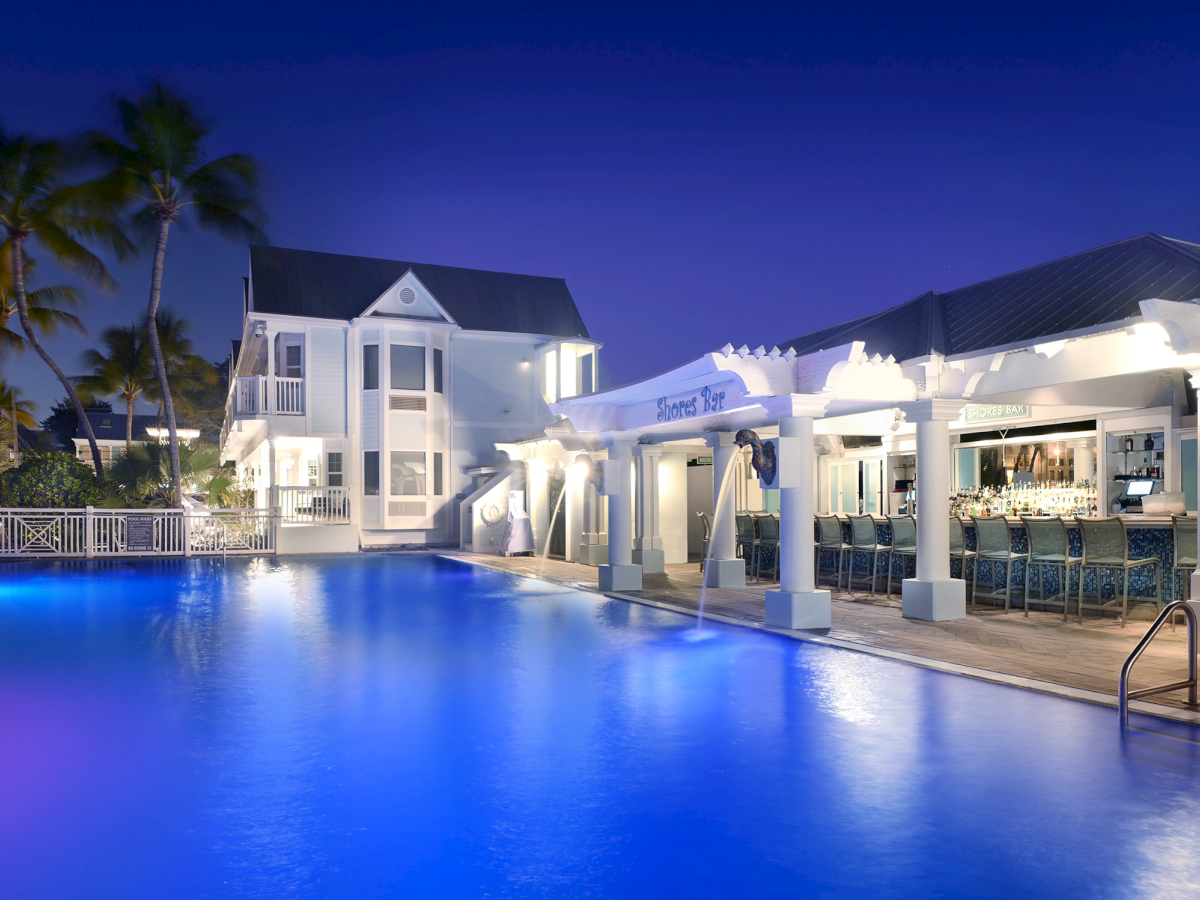 Night poolside at a resort with palm trees, white buildings, and a lit &ldquo;Shores Bar&rdquo; by the swimming pool.